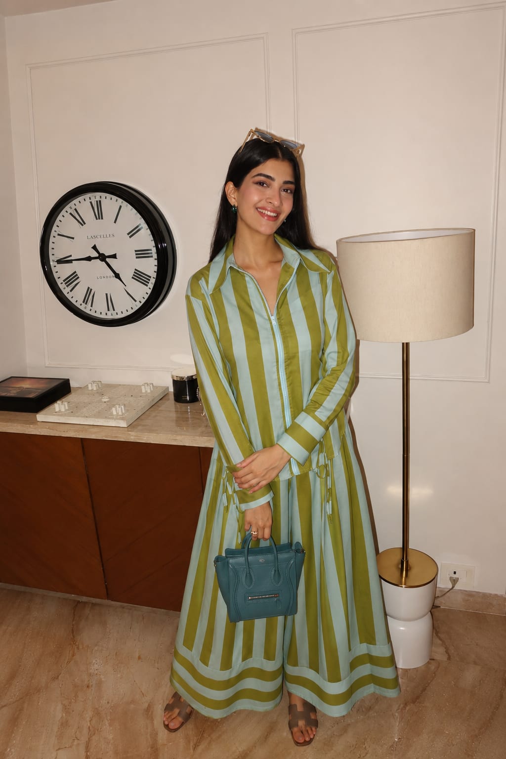 A woman wearing a green and white vertically striped long-sleeve maxi shirt dress, holding a teal blue handbag, standing in an indoor setting with a clock, lamp, and wooden cabinet in the background.