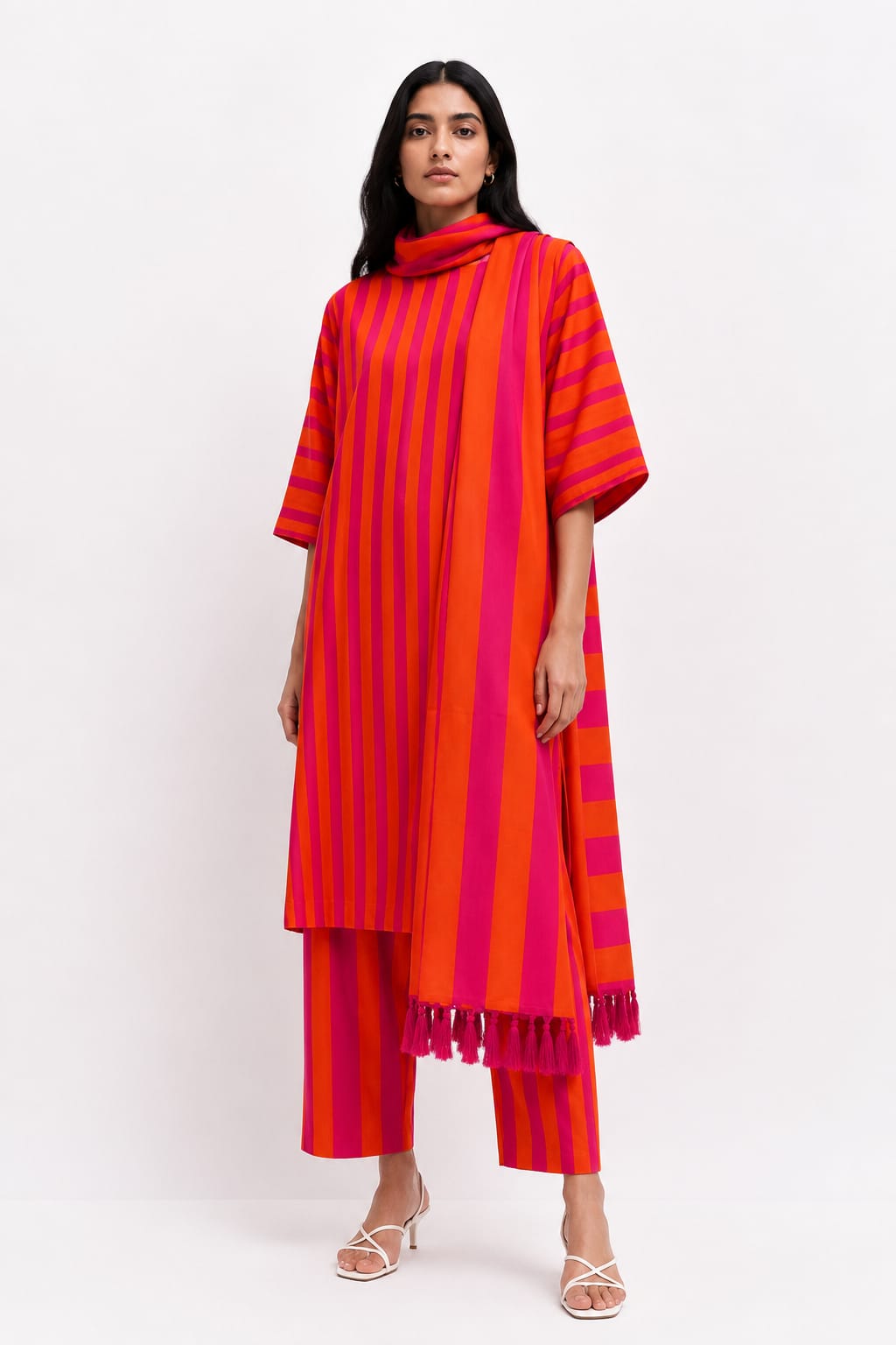 A woman modeling a vibrant red and pink striped tunic with a matching scarf and striped trousers, styled with white strappy heels against a plain white studio background.
