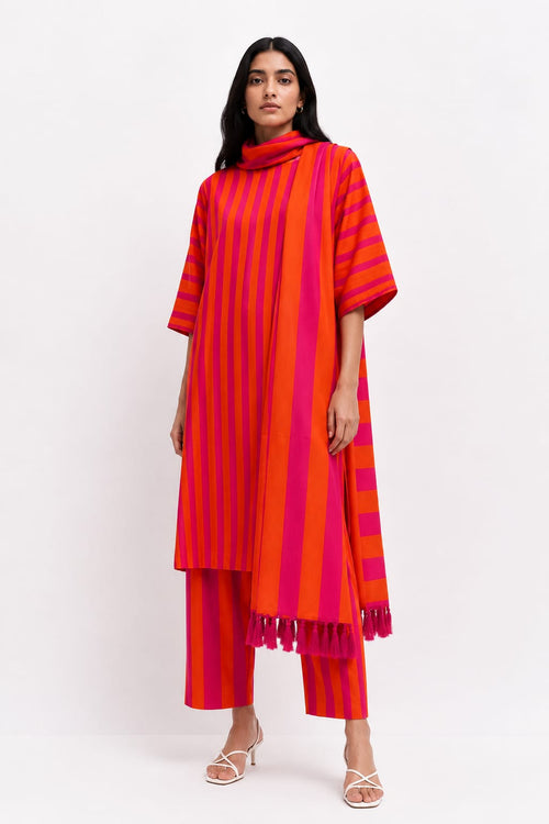 A woman modeling a vibrant red and pink striped tunic with a matching scarf and striped trousers, styled with white strappy heels against a plain white studio background.