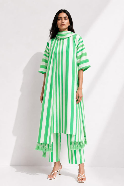 A woman modeling a green and white vertically striped tunic top with matching striped cropped pants and fringed hems, photographed against a white studio wall with soft shadows.