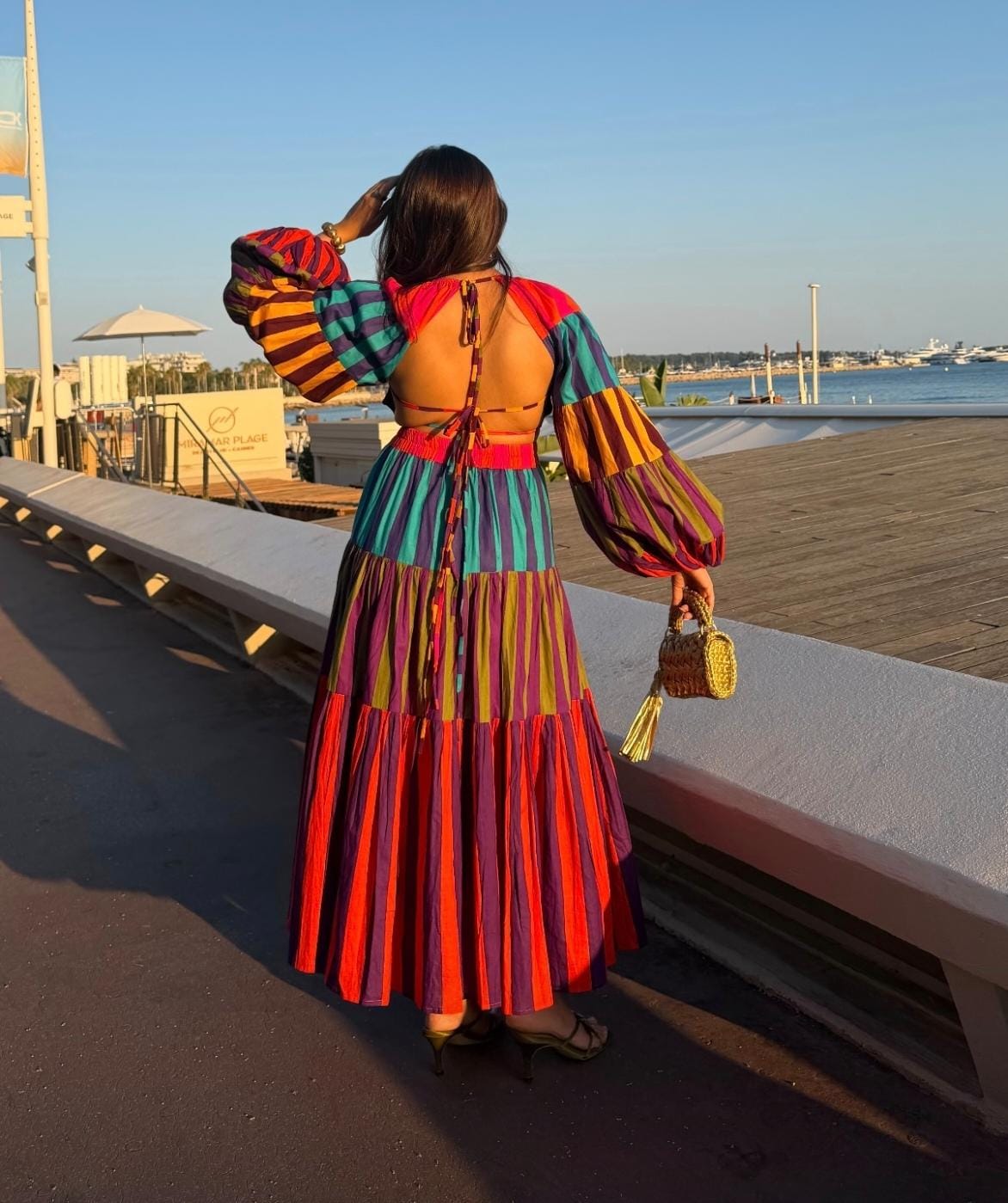A woman stands on a waterfront promenade wearing a colorful striped maxi dress with a back tie and voluminous sleeves, holding a small woven handbag.