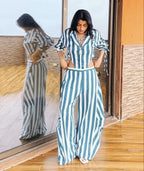 A woman stands indoors wearing a blue and white vertically striped two-piece set with a collared long-sleeve top and wide-leg pants. 