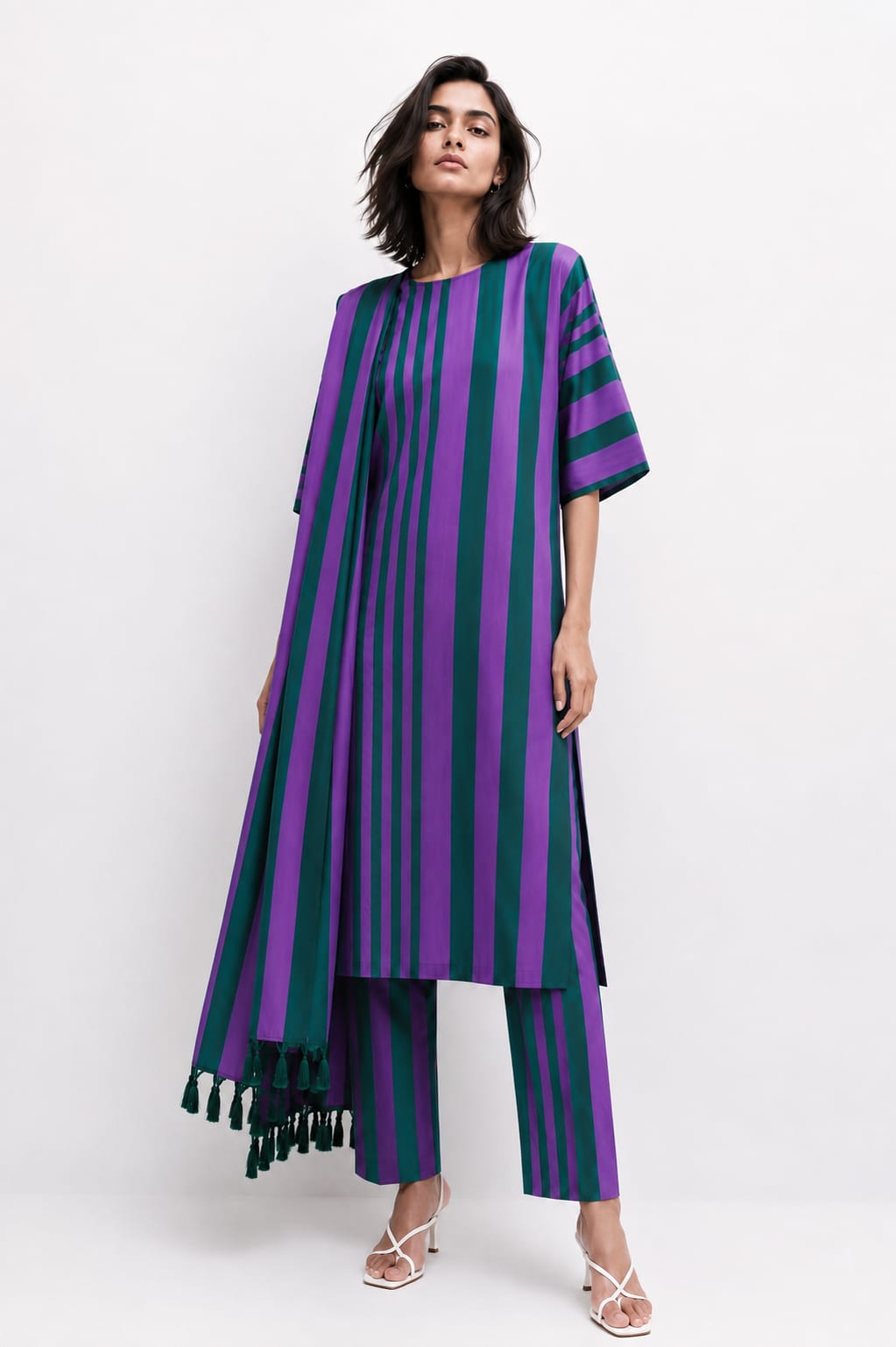 A model wearing a purple and dark green vertically striped tunic top with matching striped pants and dark green tassels, photographed against a plain white studio background.