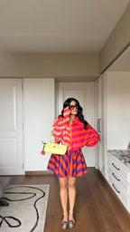 A woman stands in a home interior wearing a bright striped jacket and matching striped skirt, holding a yellow handbag. 
