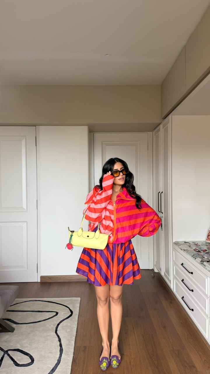 A woman stands in a home interior wearing a bright striped jacket and matching striped skirt, holding a yellow handbag. 