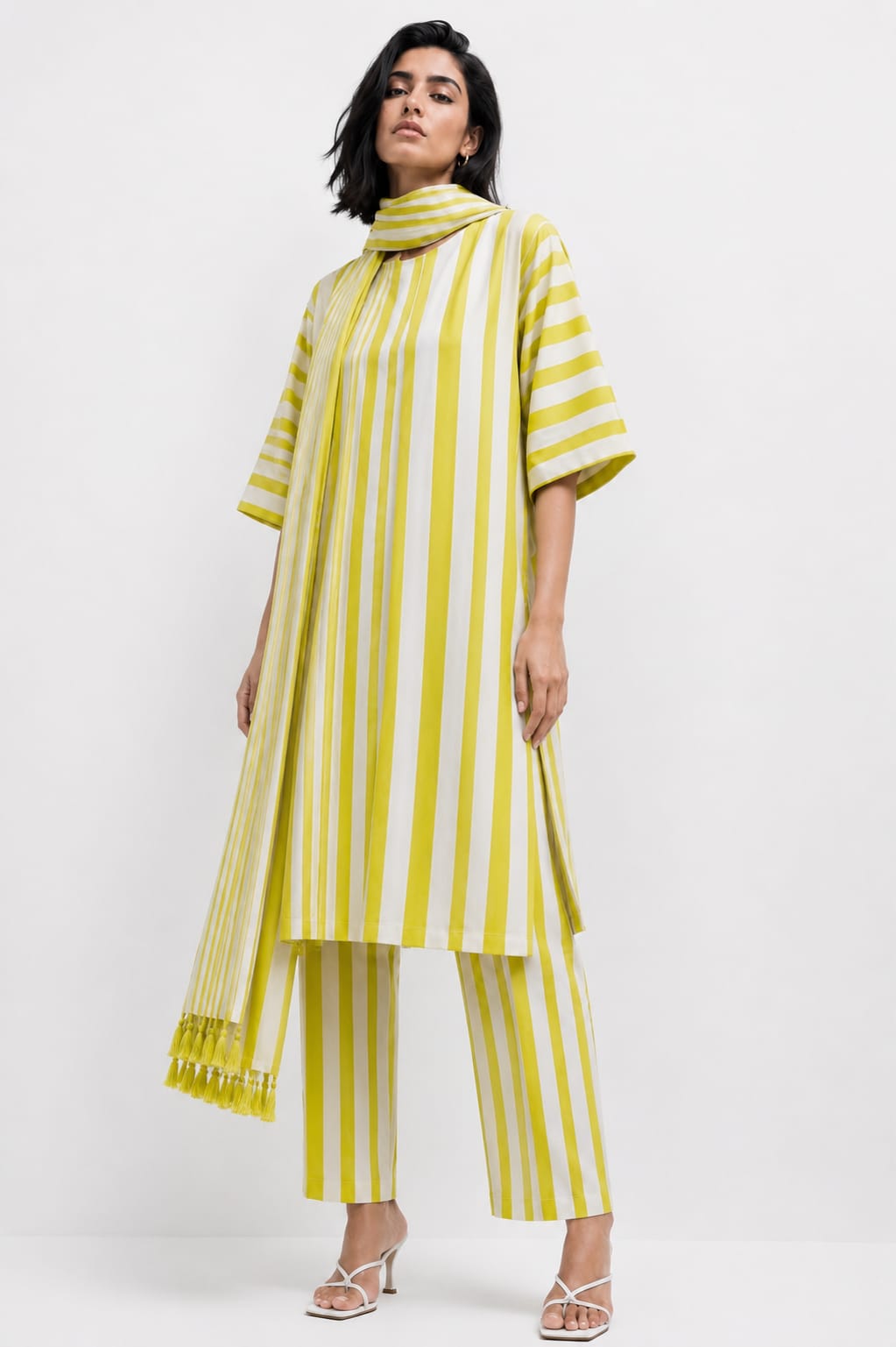 A model wearing a yellow and white vertically striped tunic top with matching striped pants and white heeled sandals, photographed against a plain white studio background.