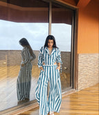 A woman wearing a blue and white vertically striped matching set stands on a wooden deck in front of large glass doors, with her reflection visible in the mirror. The outfit includes a collared long-sleeve top and wide-leg trousers.