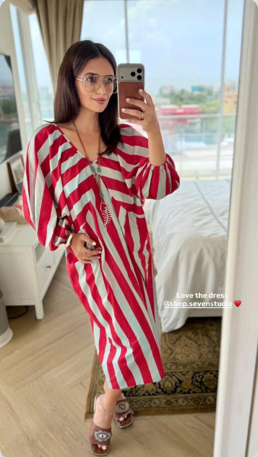 A woman takes a mirror selfie in a hotel room wearing a red and white vertically striped maxi dress with wide sleeves and a V-neckline.