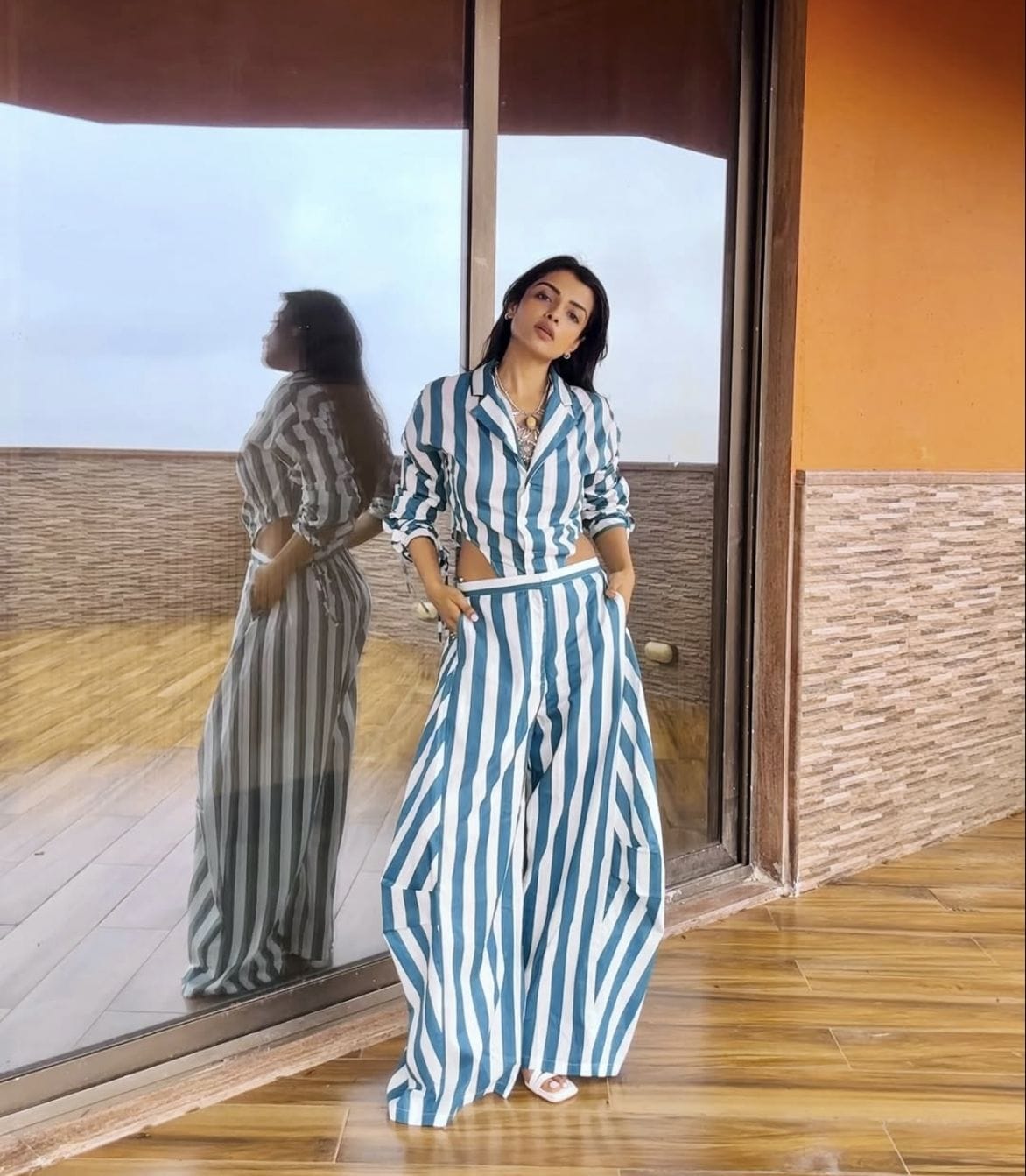 A woman stands indoors by a large glass window wearing a blue and white vertically striped button-up shirt and matching wide-leg pants. Her reflection is visible in the glass, and the room has wood flooring and an orange-brown wall.